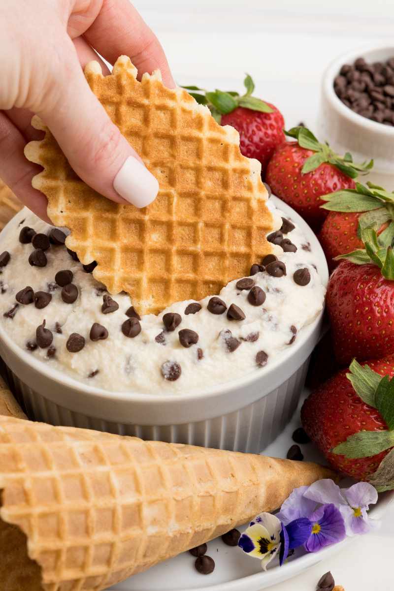 A hand dips a waffle cookie into a large ramekin of cannoli dip garnished with mini chocolate chips.