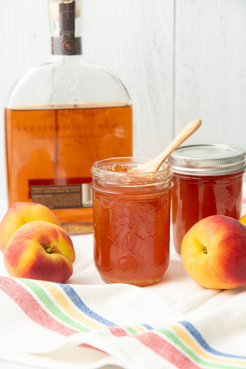 A bottle of bourbon, two jars of homemade jam, and three fresh peaches arranged on a striped kitchen linen.