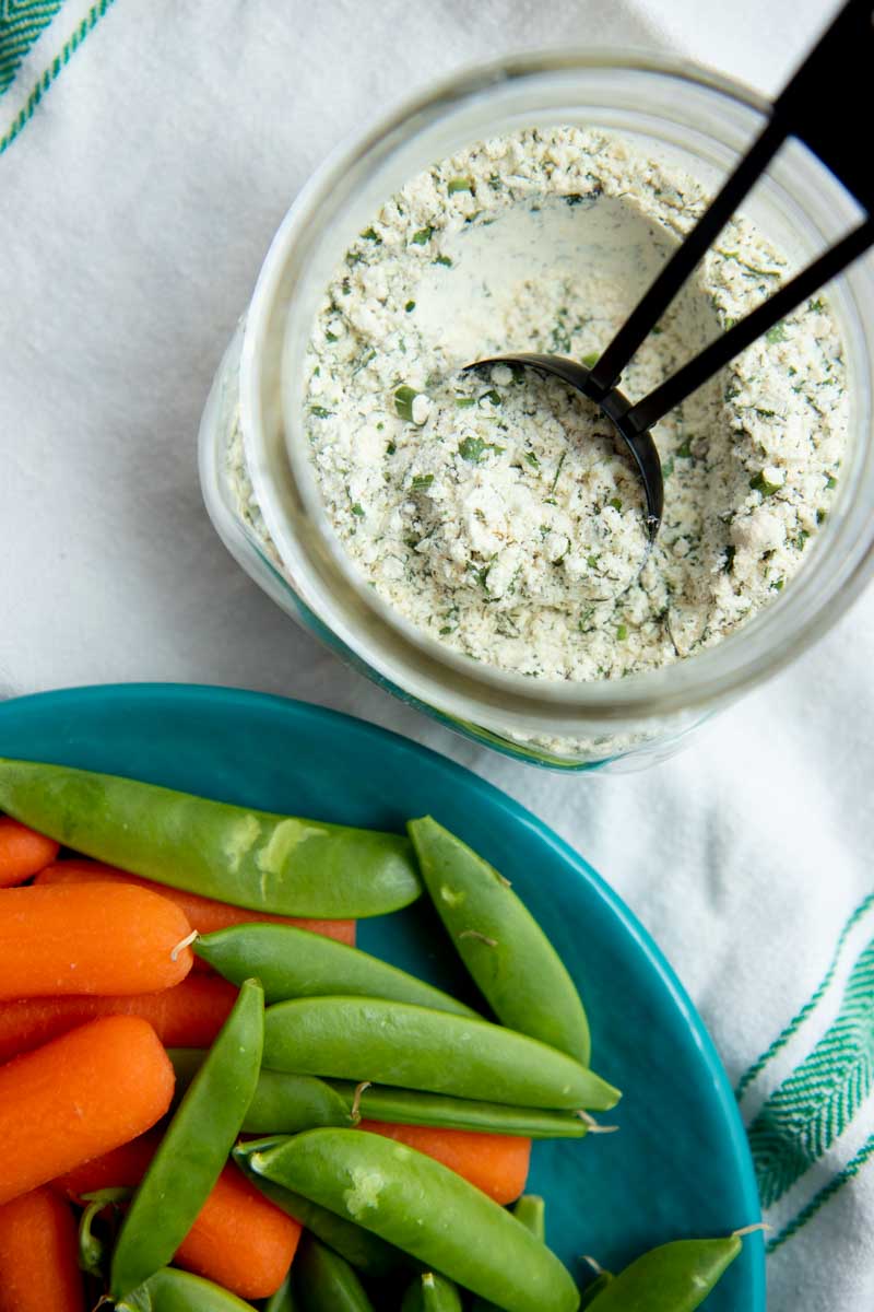 Overhead of an open jar of ranch seasoning mix with a scoop in it with a plate of baby carrots and snap peas alongside.