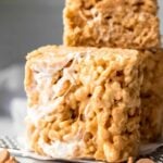A peanut butter and marshmallow cereal square stands on a parchment paper lined cooling rack with three other cereal treats stacked behind it.