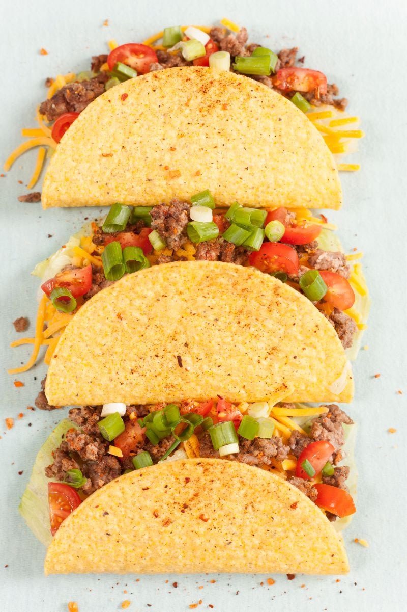 A trio of crunchy tacos stuffed with meat, cheese, lettuce, and tomatoes on a light background.