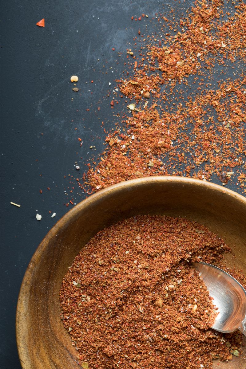 A wooden bowl filled with gluten-free taco seasoning rests on a counter with extra seasoning around the bowl.