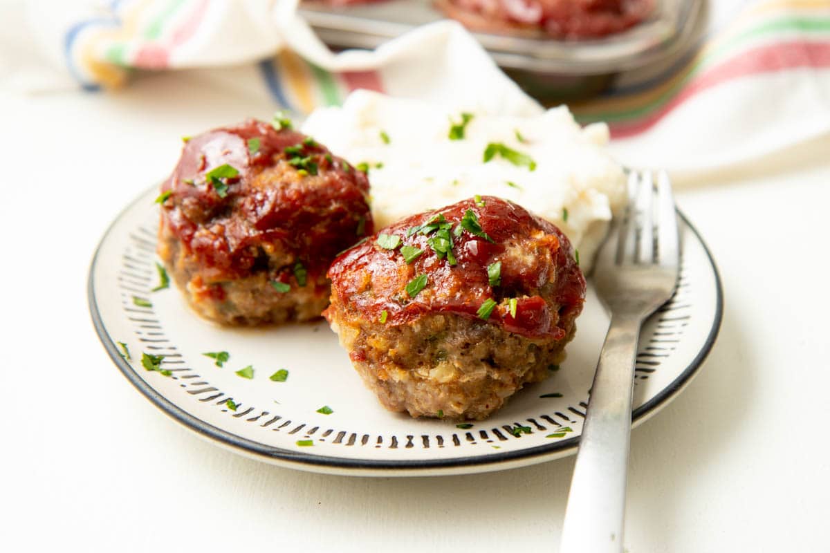 A glazed mini meatloaf is served on a white plate with a side of mashed potatoes and a second meatloaf.