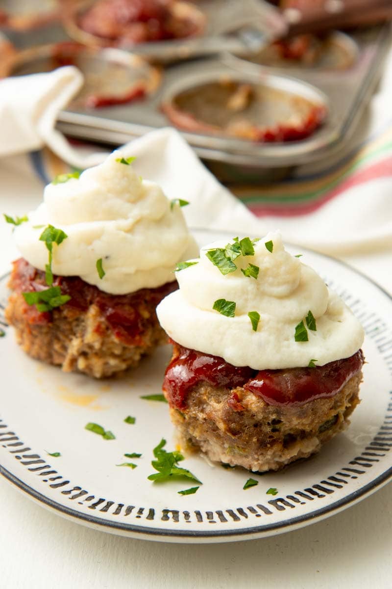 A glazed mini meatloaf topped with mashed potatoes rests on a plate, a second mini meatloaf beside it.