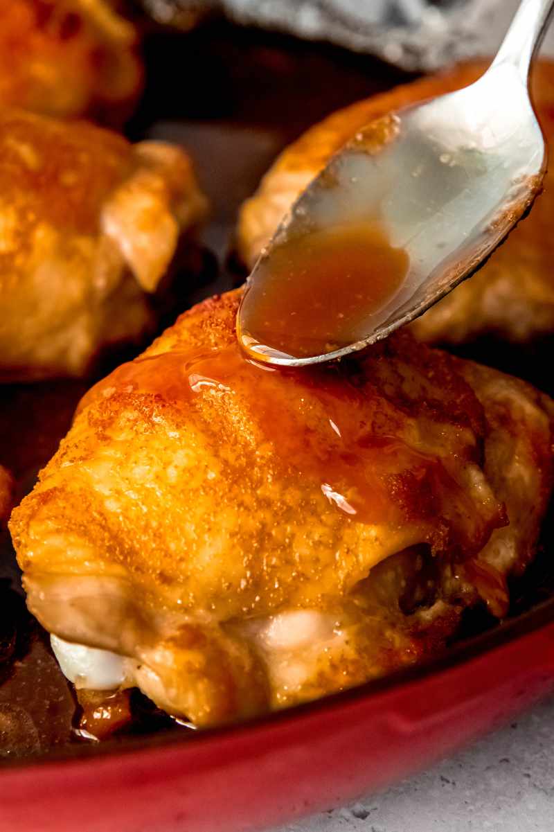Close view of a spoon basting sauce over a baked chicken thigh in an enameled cast iron skillet.