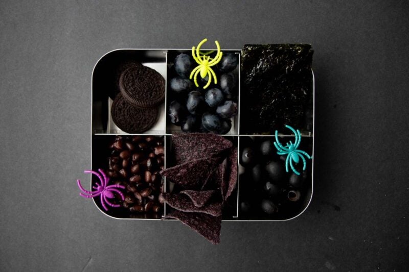Overhead of a six-compartment bento-style lunchbox filled with all black foods and colorful plastic spiders.