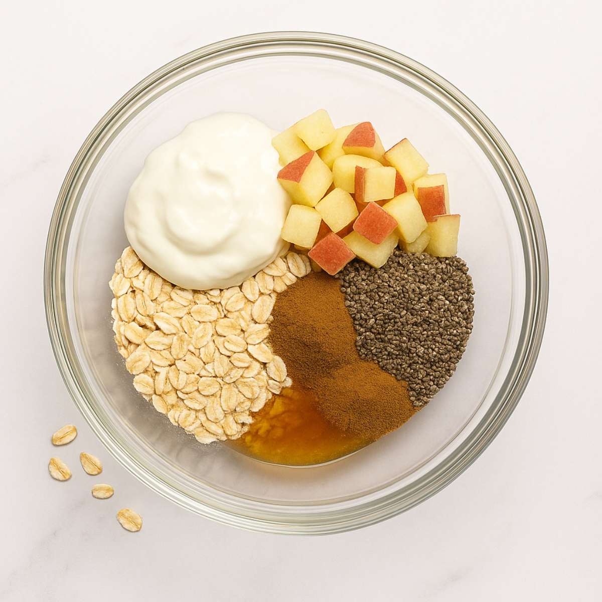 Ingredients for apple cinnamon overnight oats in a glass bowl on a white marble countertop