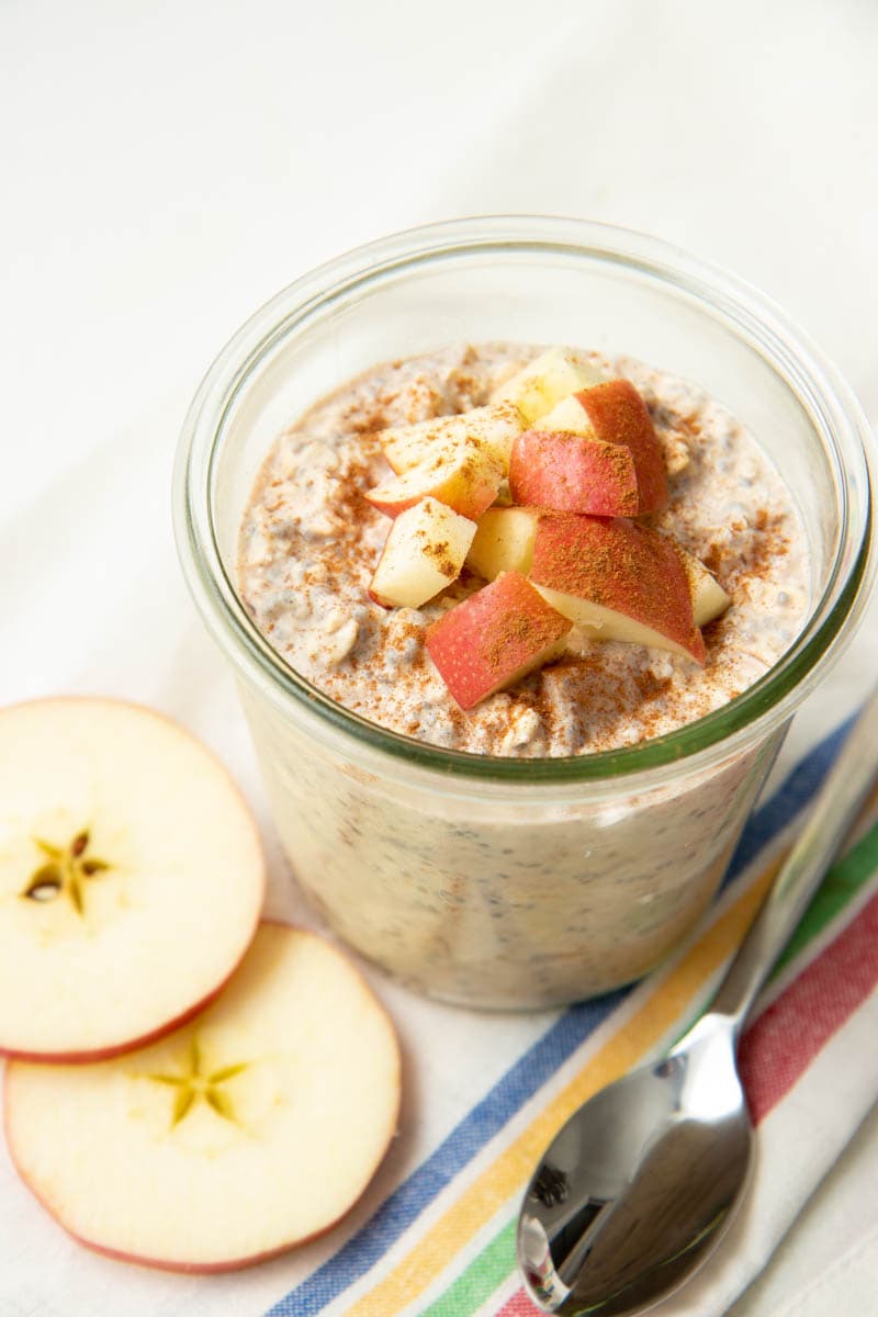A jar of apple cinnamon overnight oats topped with chopped apples and cinnamon rests on a kitchen linen with a spoon and apple slices beside it.