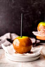 A homemade caramel apple rests on a square of parchment paper atop a small stack of plates.