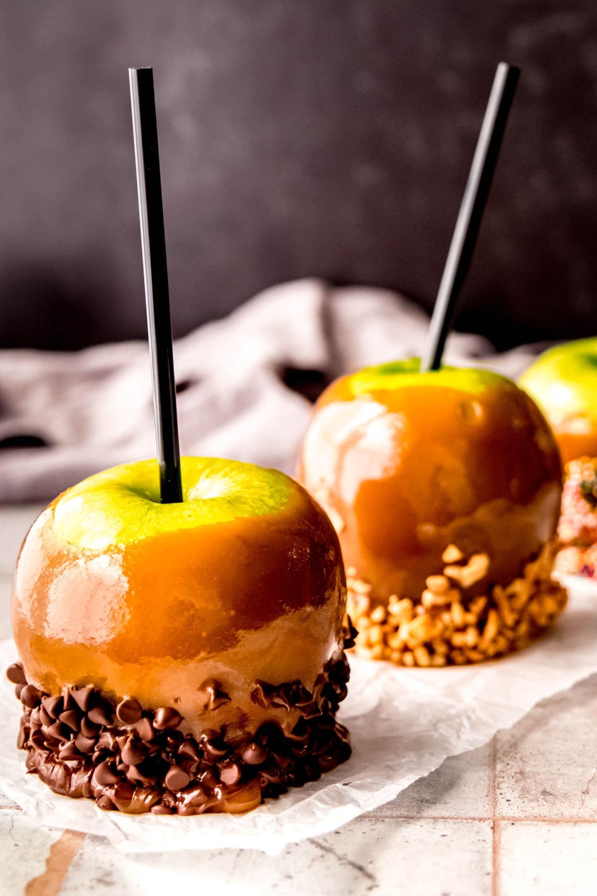 Three caramel apples sit on a piece of parchment paper on a countertop