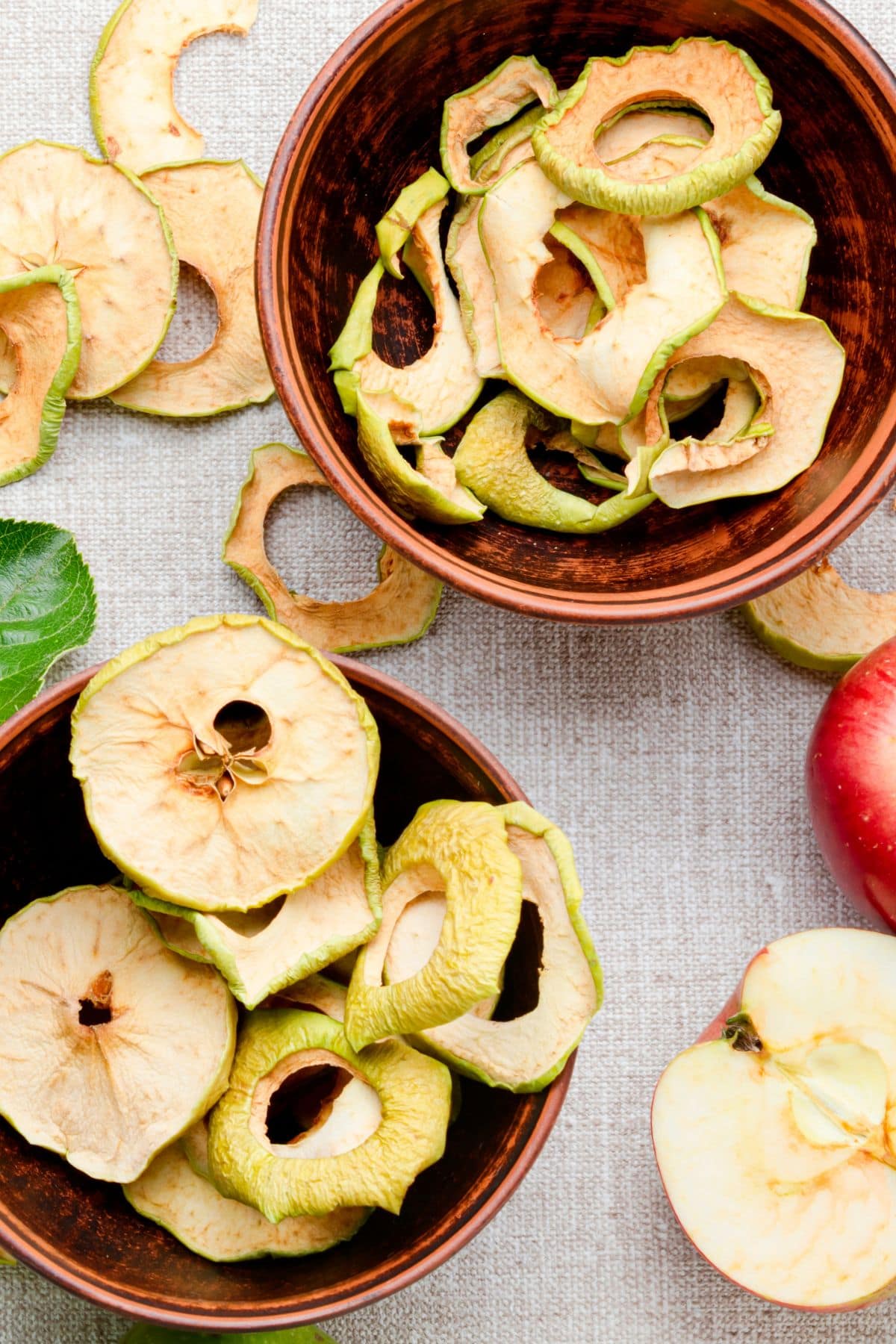 Two bowls of dried apple slices sit on a table with apples around them.