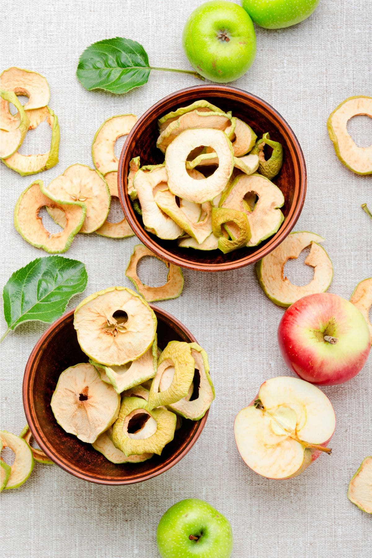 Two bowls of dried apple slices sit on a table with apples around them.