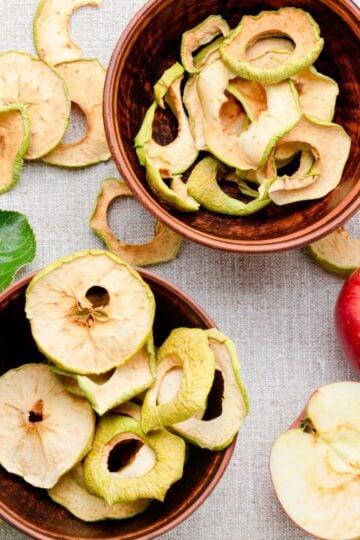 Two bowls of dried apple slices sit on a table with apples around them.