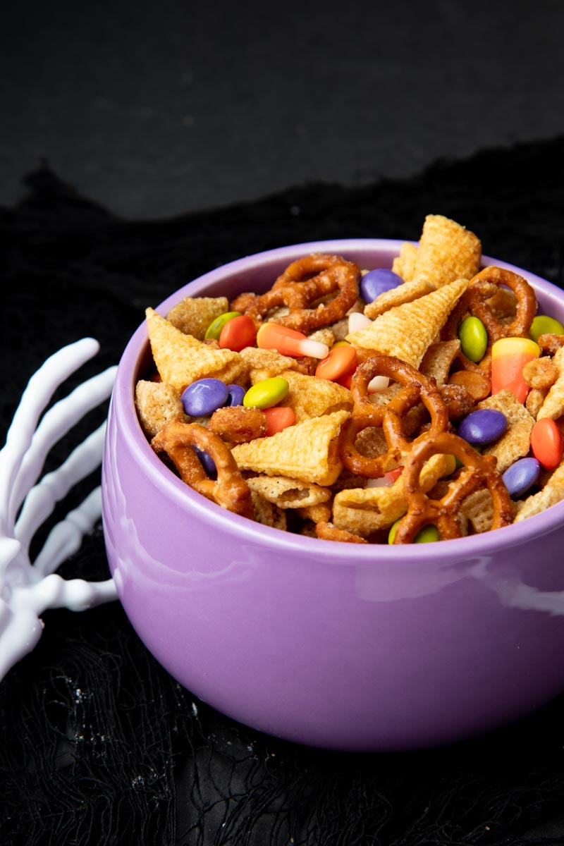 A skeleton hand reaches for a purple bowl filled to the brim with Halloween chex mix.