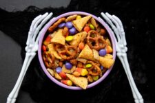 Two skeleton arms reach out, bony fingers curving around a bowl of Halloween chex mix with pretzels, bugles, and M&Ms.