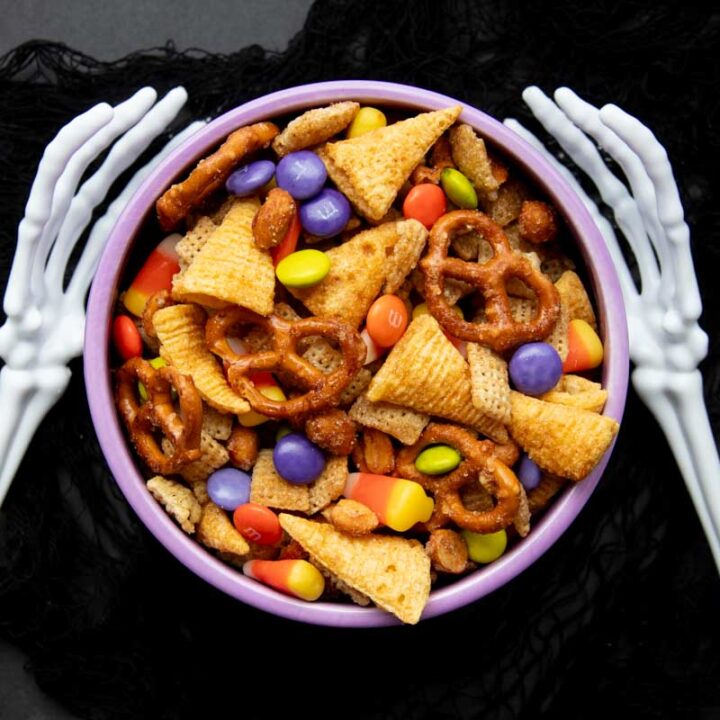 Two skeleton arms reach out, bony fingers curving around a bowl of Halloween chex mix with pretzels, bugles, and M&Ms.