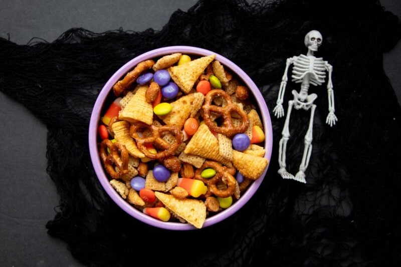 Overhead of a purple serving bowl filled with Halloween chex mix on black fabric with a plastic skeleton nearby.