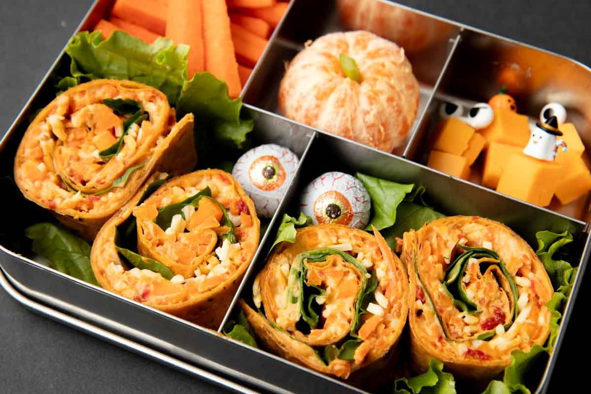 Halloween veggie pinwheels nestles into a bento-style lunchbox with other orange foods.