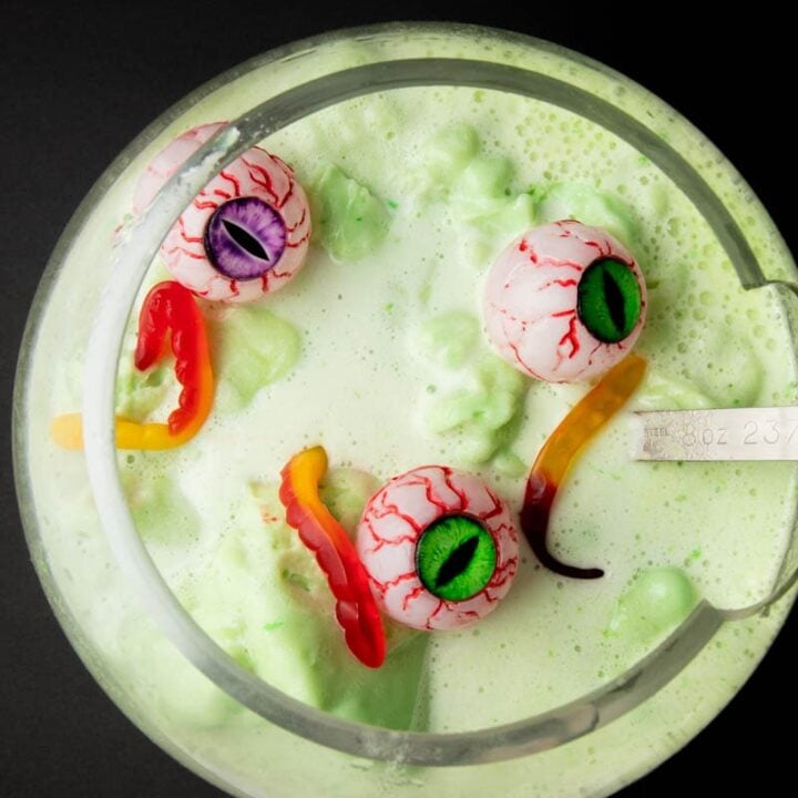 Overhead of a ladle in a large punch bowl filled with Halloween punch garnished with gummy worms and plastic eyes on a black background.