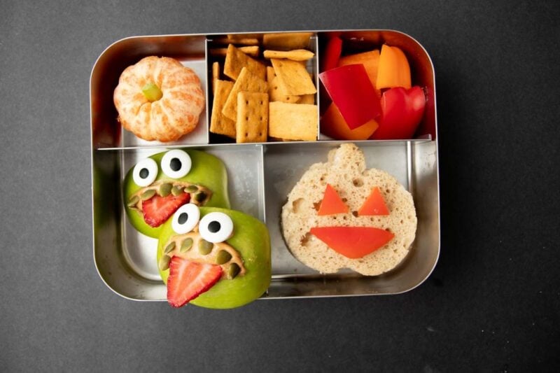 Top view of a Halloween lunch idea: a sandwich bento with monster apple snacks, clementine pumpkin, cheddar crackers, and red and orange bell pepper pieces.
