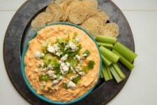 Overhead of a serving platter with a teal bowl of Instant Pot buffalo chicken dip surrounded by round tortilla chips and celery sticks.