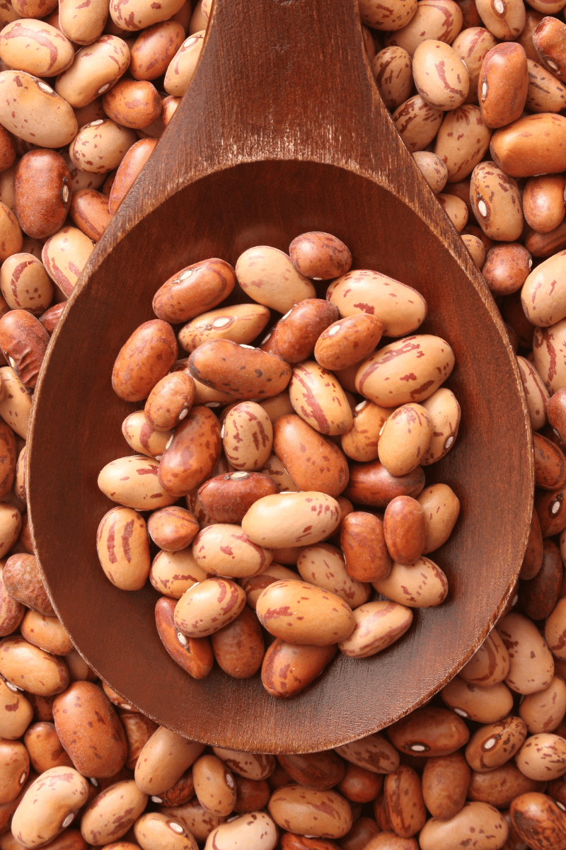 Dried pinto beans rest in the bowl of a large wooden spoon atop a layer of more dried pintos.