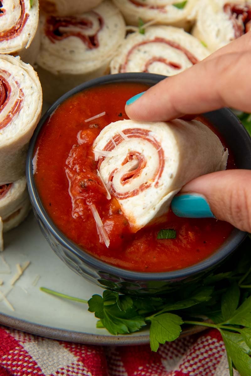 A hand dunks a roll-up slice into a small bowl of marinara sauce on a platter of roll-ups.