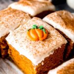 An edible pumpkin garnish rests atop the cream cheese frosting on a piece of cake dusted with cinnamon.