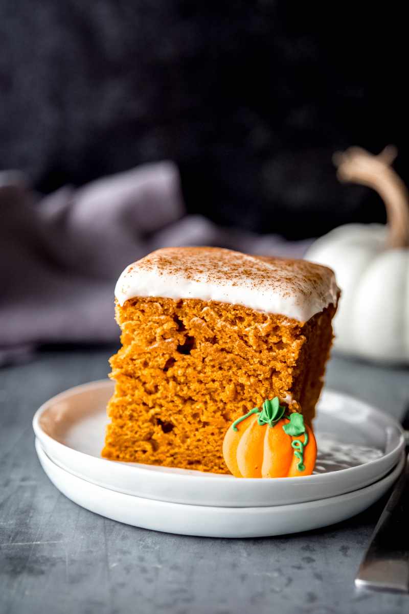 A piece of pumpkin cake with cream cheese frosting on a white dish with an edible pumpkin garnish.