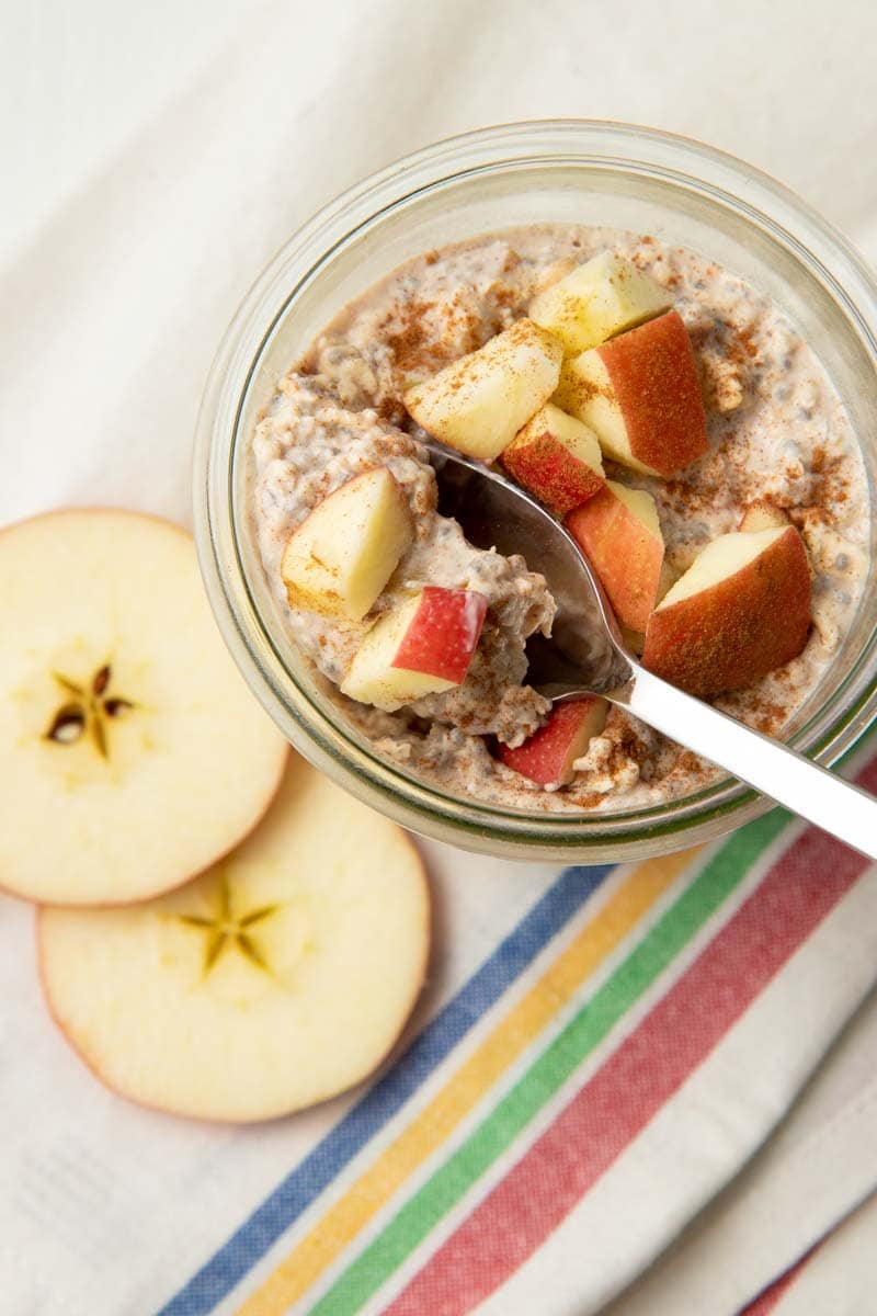 A spoon scoops up apple overnight oats from the top of a glass jar.