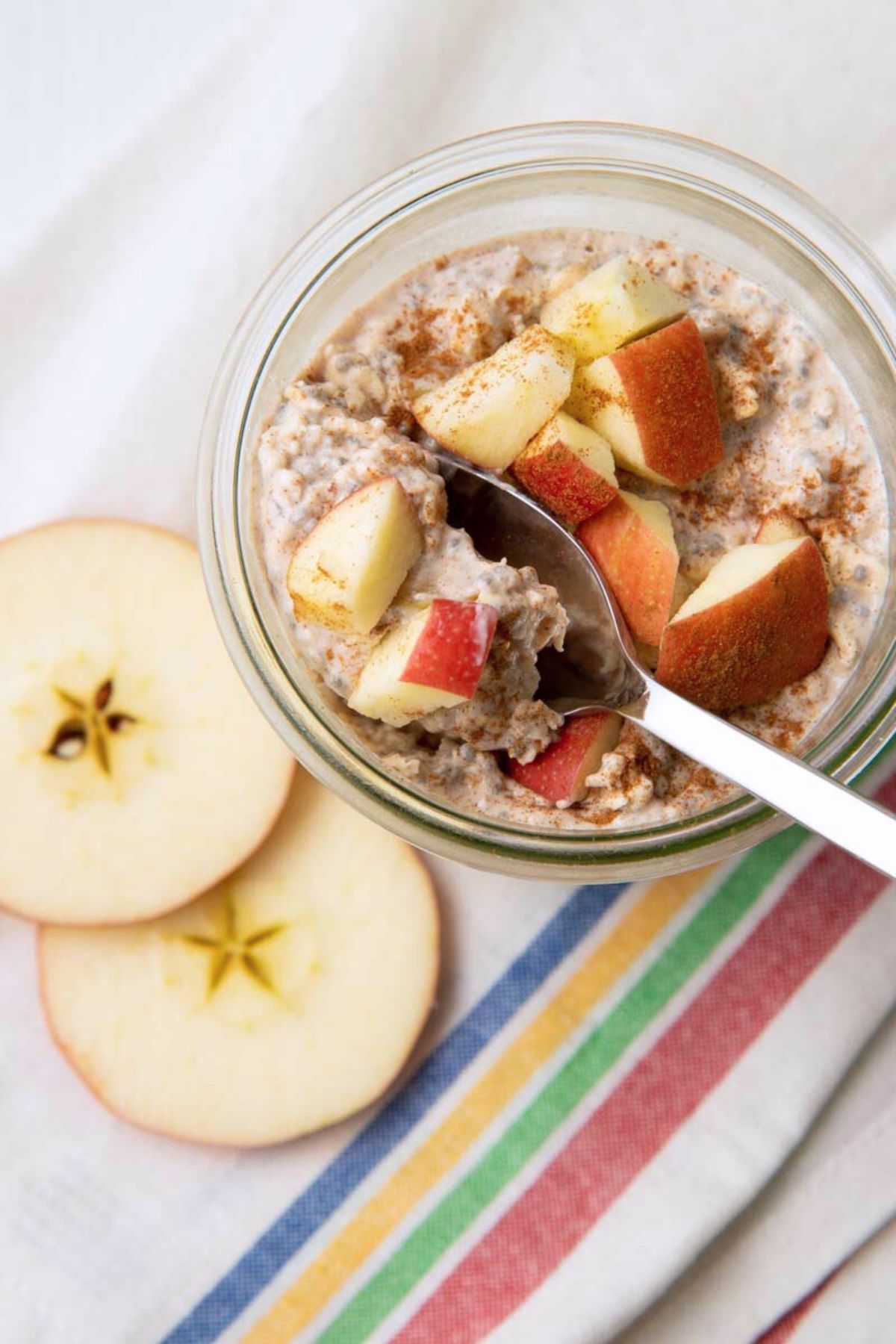 A spoon scoops out a bite from a jar of apple overnight oats sitting on top of a striped kitchen towel