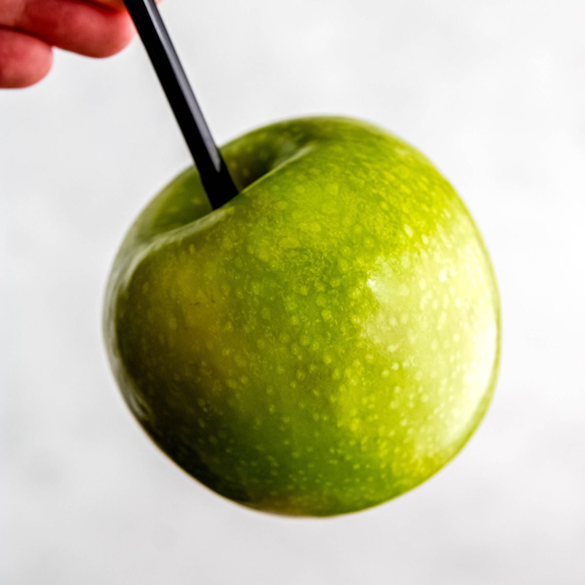 A hand holds a green apple with a black stick inserted into it