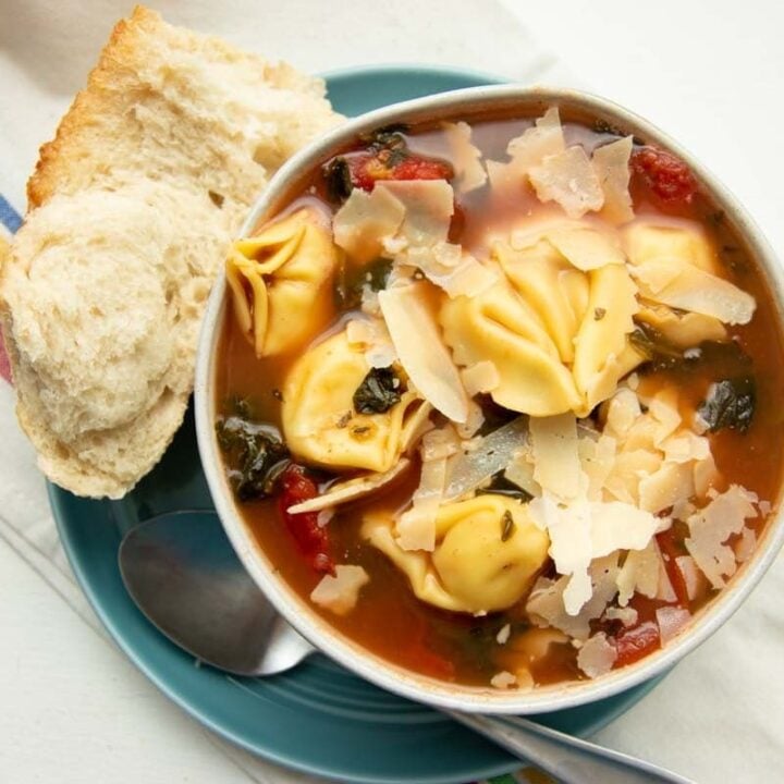 A bowl of tortellini soup garnished with shaved parmesan rests on a plate atop a striped kitchen linen.