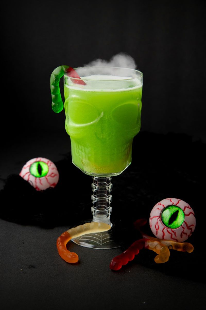A glass of spooky green witches' brew in a skull-shaped goblet with gummy works and plastic bloodshot eyes around the stem.