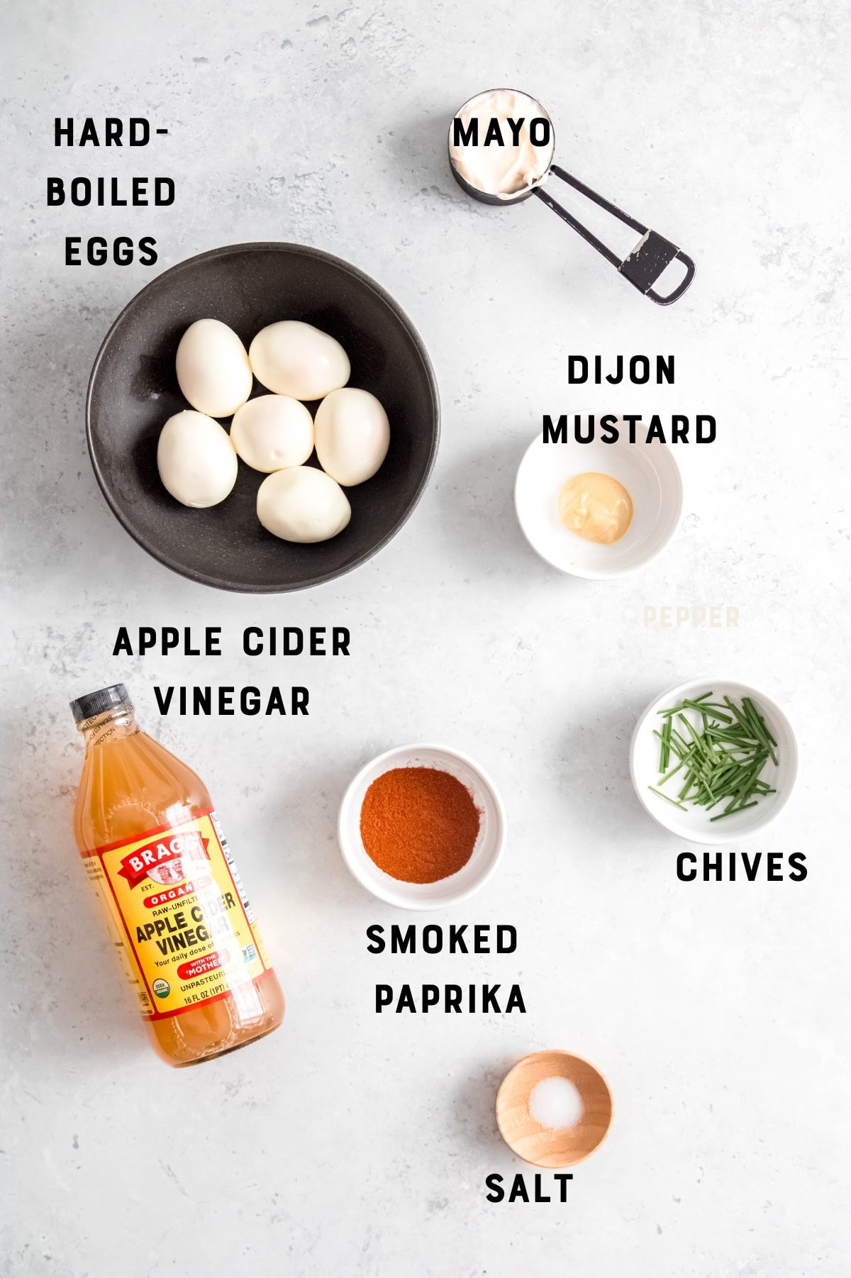 Ingredients for deviled eggs sit on a white countertop: hard-boiled eggs, mayonnaise, dijon mustard, chives, smoked paprika, apple cider vinegar, salt.