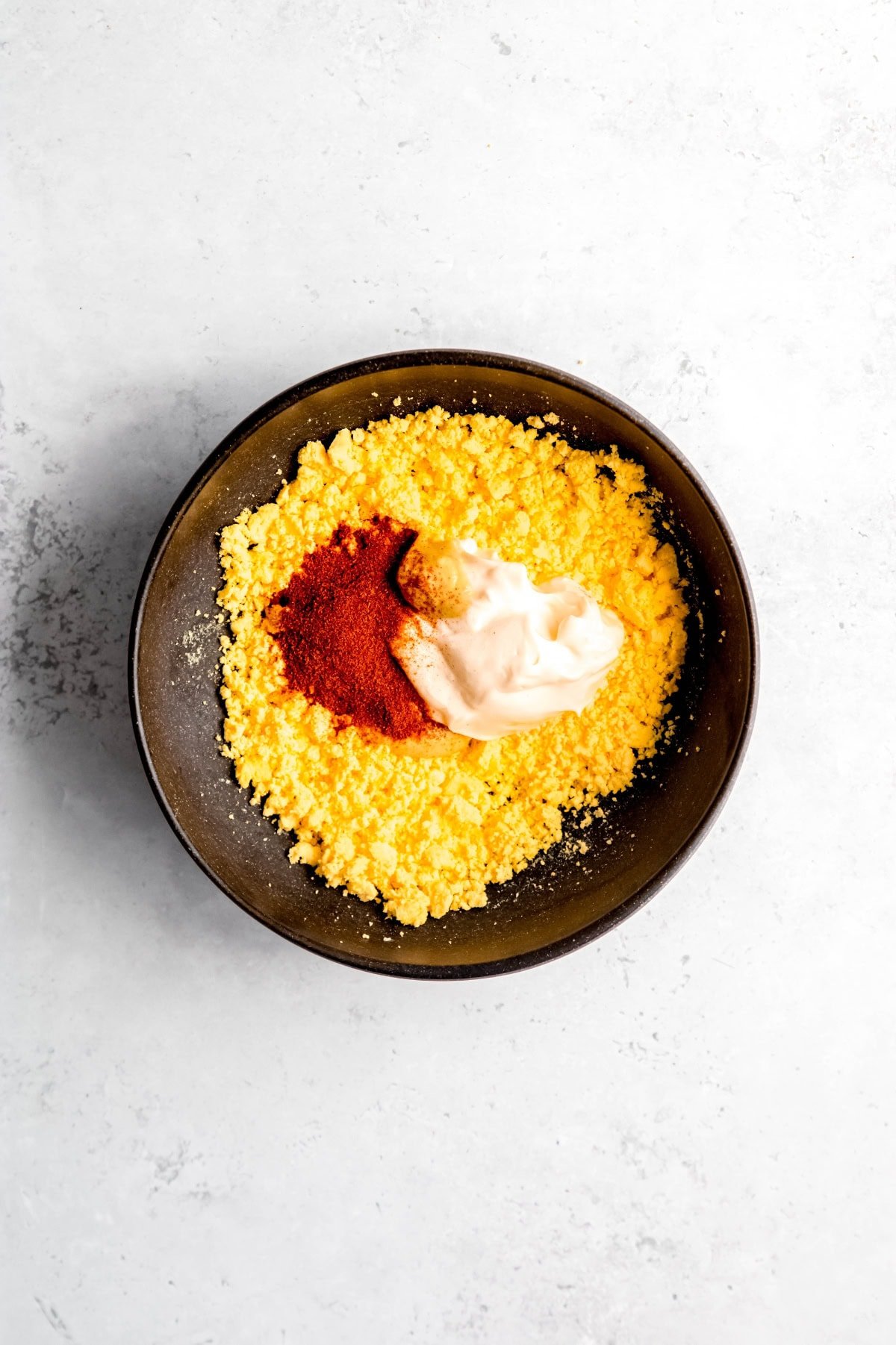 Mayonnaise, dijon mustard, and spices on top of mashed hard-boiled egg yolks in a dark bowl.