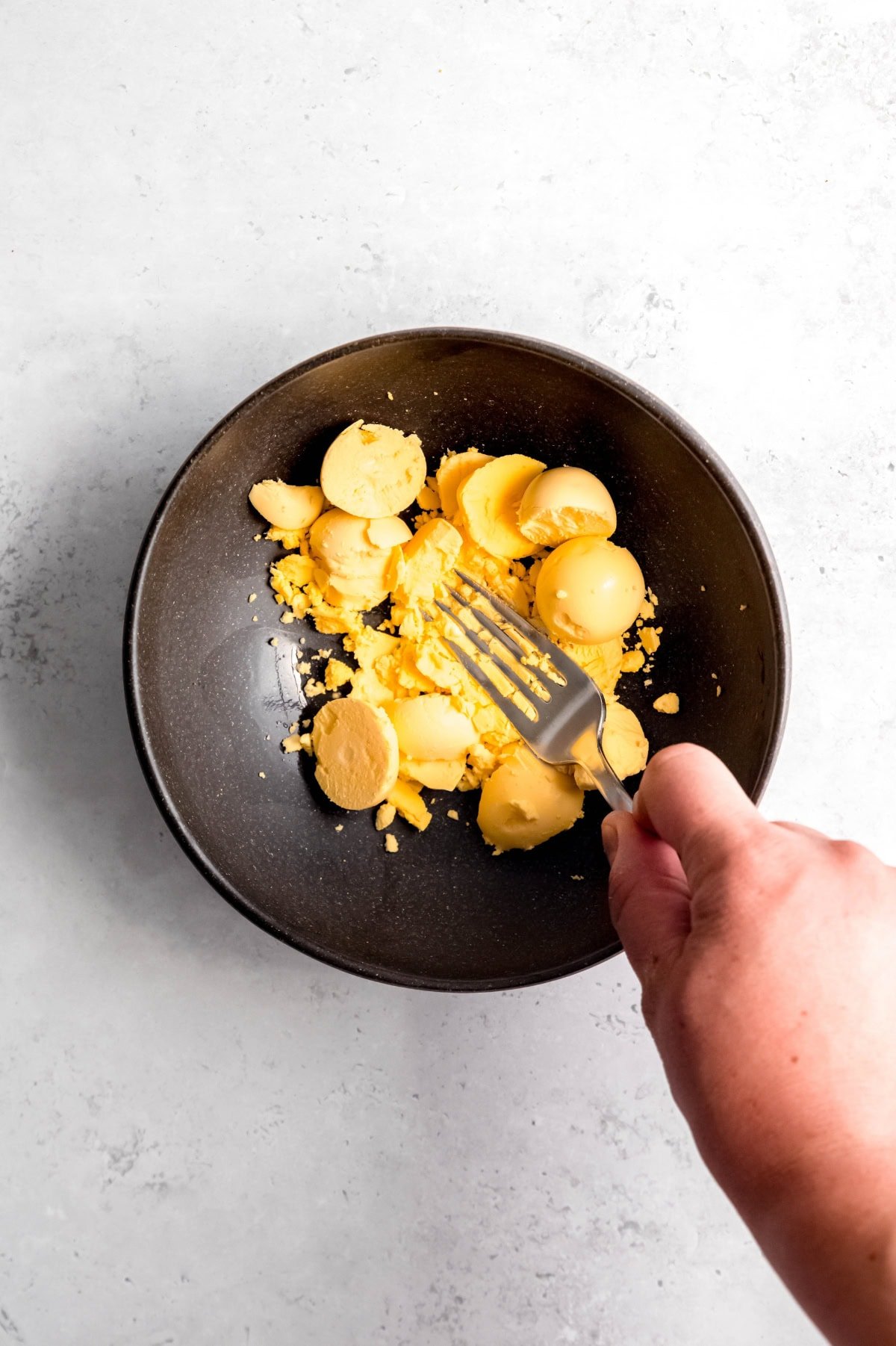 A hand uses a fork to mash hard-boiled egg yolks in a dark bowl.