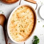 Overhead of a creamy potato casserole in a white baking dish on a light counter with a wooden spoon alongside.