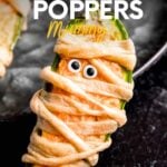 Close view of a mummy jalapeno popper standing up against the edge of a serving platter. A text overlay reads, "Jalapeno Poppers Mummy!"