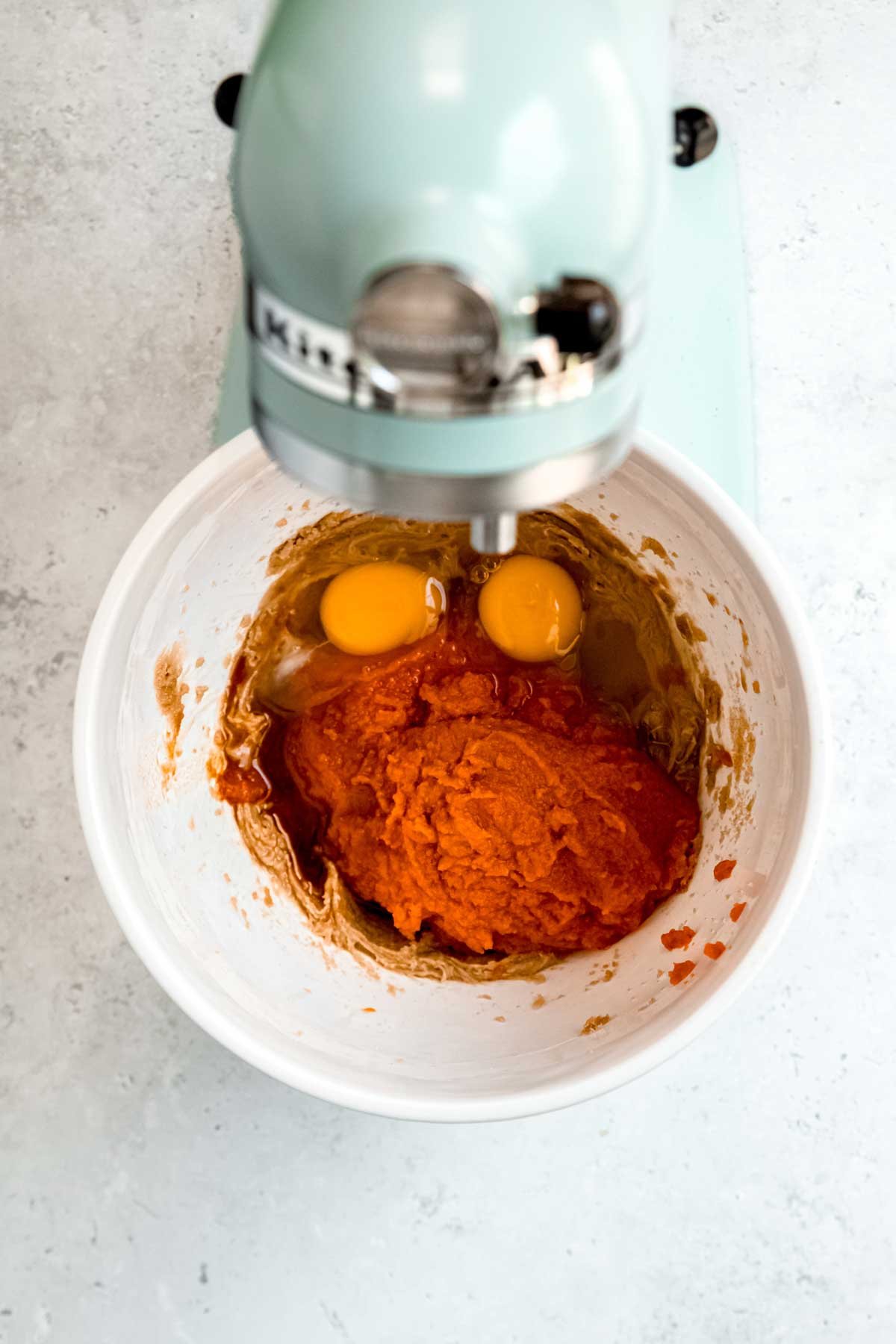 A blue stand mixer blends together eggs, pumpkin puree, and other wet cake ingredients in a white bowl