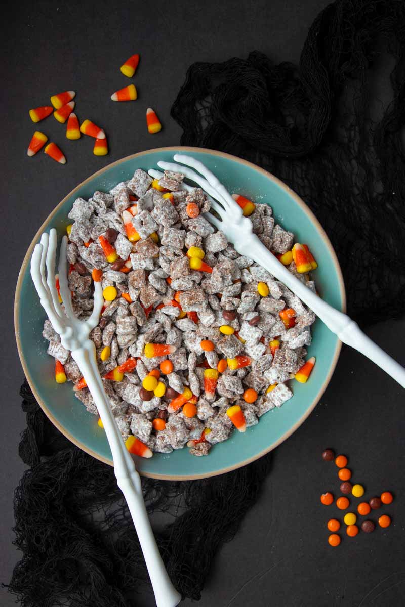 Two long skeleton arms extend into a bowl of puppy chow with candy corn and Reese's Pieces.