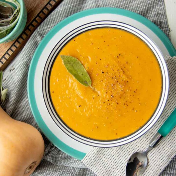 A serving of roasted squash soup garnished with a sage leaf in a bowl on a platter with a spoon and cloth napkin alongside.