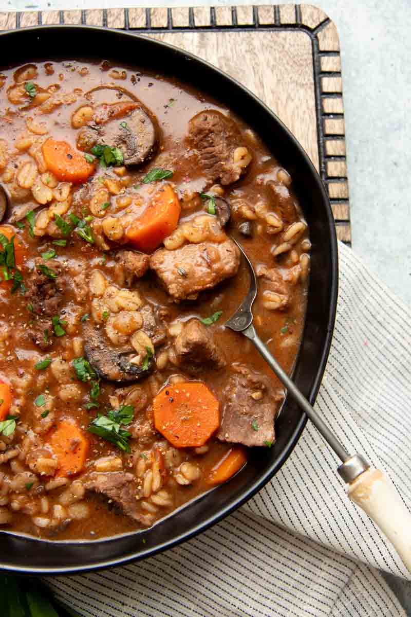 Overhead of a long metal spoon with a wooden handle scooping into a bowl of rich beef soup.