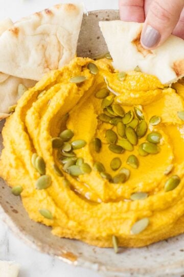 Scooping pumpkin hummus out of a bowl with a pita wedge