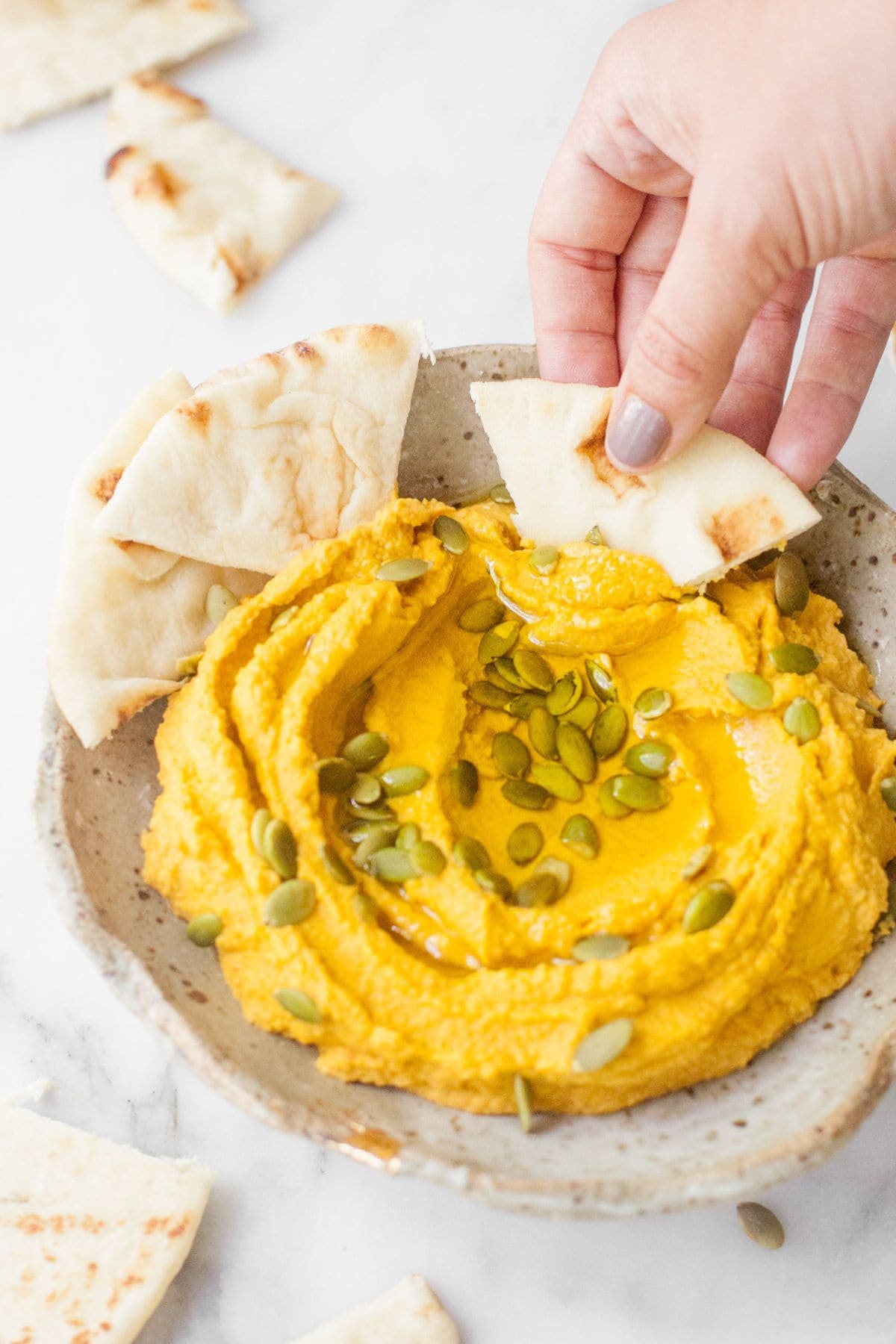 A hand scoops a pita wedge into a bowl of pumpkin hummus
