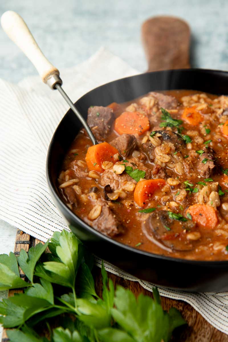 A steaming bowl of beef barley soup in a large black serving bowl on a kitchen linen with fresh sprigs of flatleaf parsley nearby.