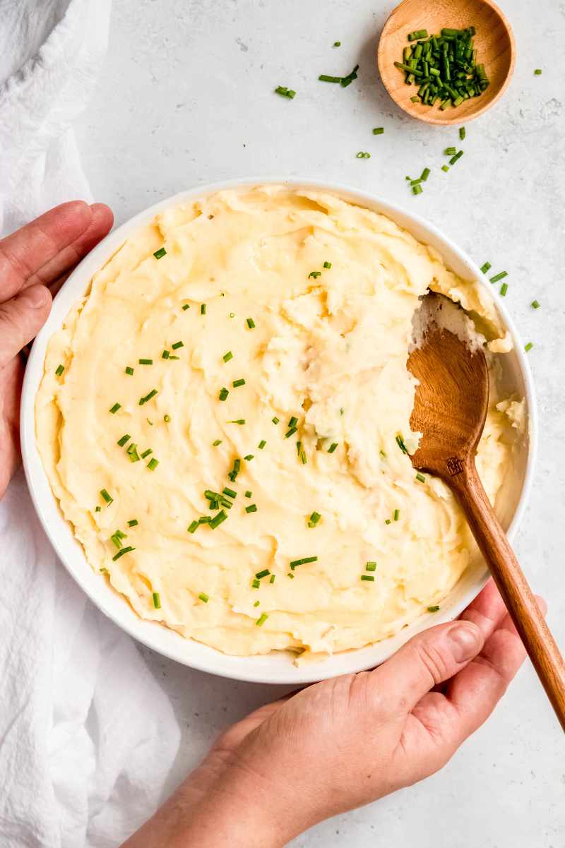 Two hands set down a serving bowl of creamy mashed potatoes on a light counter.