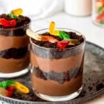 Two single-serving dirt cakes on a tray with additional dirt crumbs and gummy worms around.
