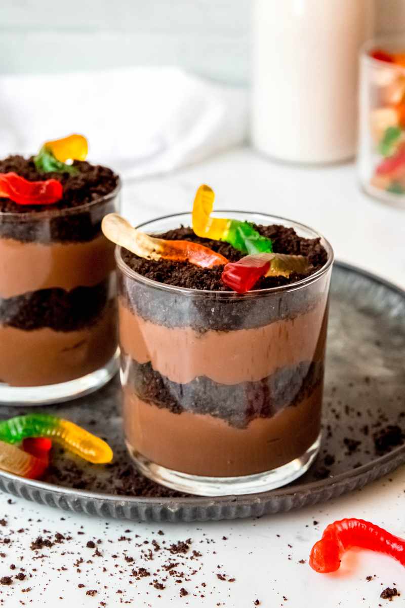 Two single-serving dirt cakes on a tray with additional dirt crumbs and gummy worms around.