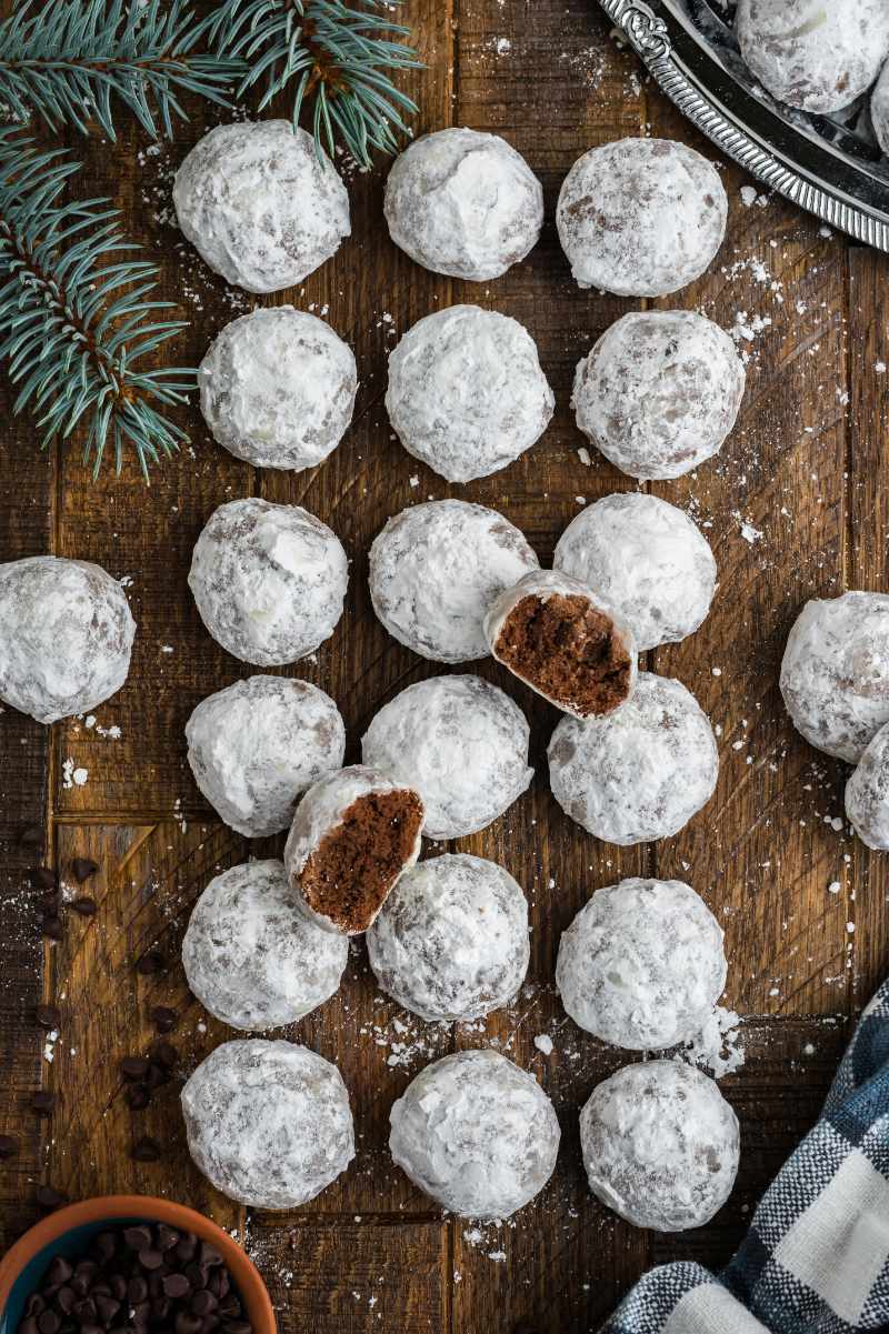 Overhead of chocolate snowball cookies in three long rows on a wooden table, additional powdered sugar and chocolate chips around.